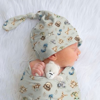 A sleeping newborn wrapped in a swaddle with a sage coloured top knot beanie on the head, featuring a pattern with various australian animals.