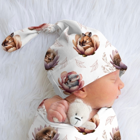 Baby wearing a top knot beanie with animal prints and holding a teddy bear on a soft white background
