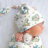 Baby wearing a dinosaur-themed top knot beanie and blanket on a white background