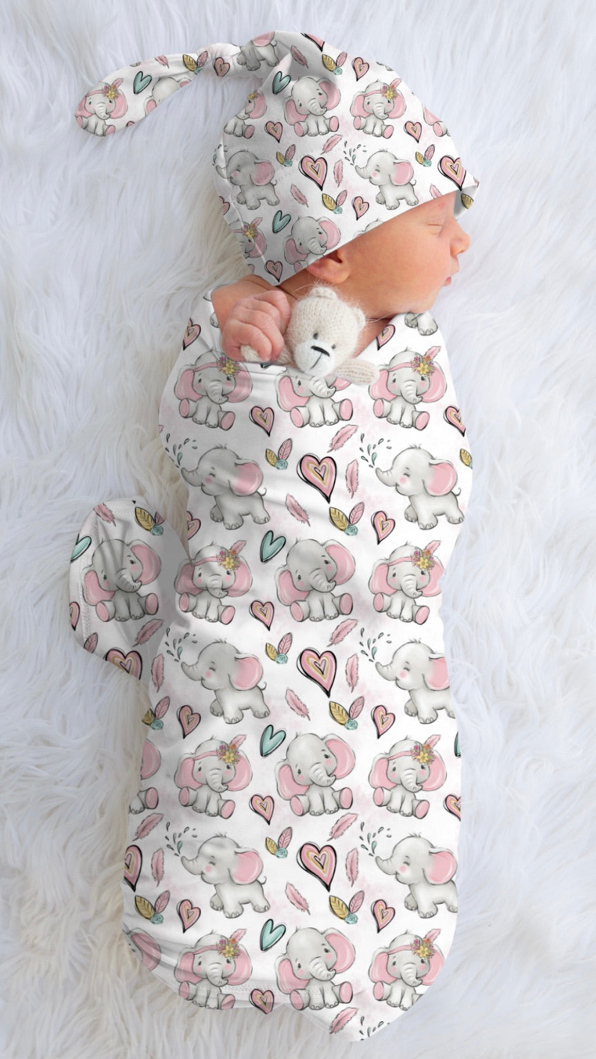 A baby swaddle wrap with a pink elephant and hearts pattern, displayed on a white fluffy surface.