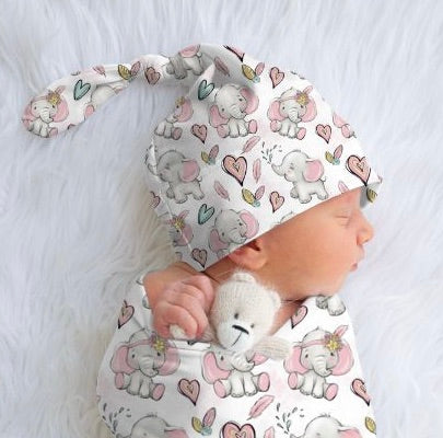A newborn baby wearing a pink top knot beanie with elephant and hearts pattern, lying on a white fluffy surface.